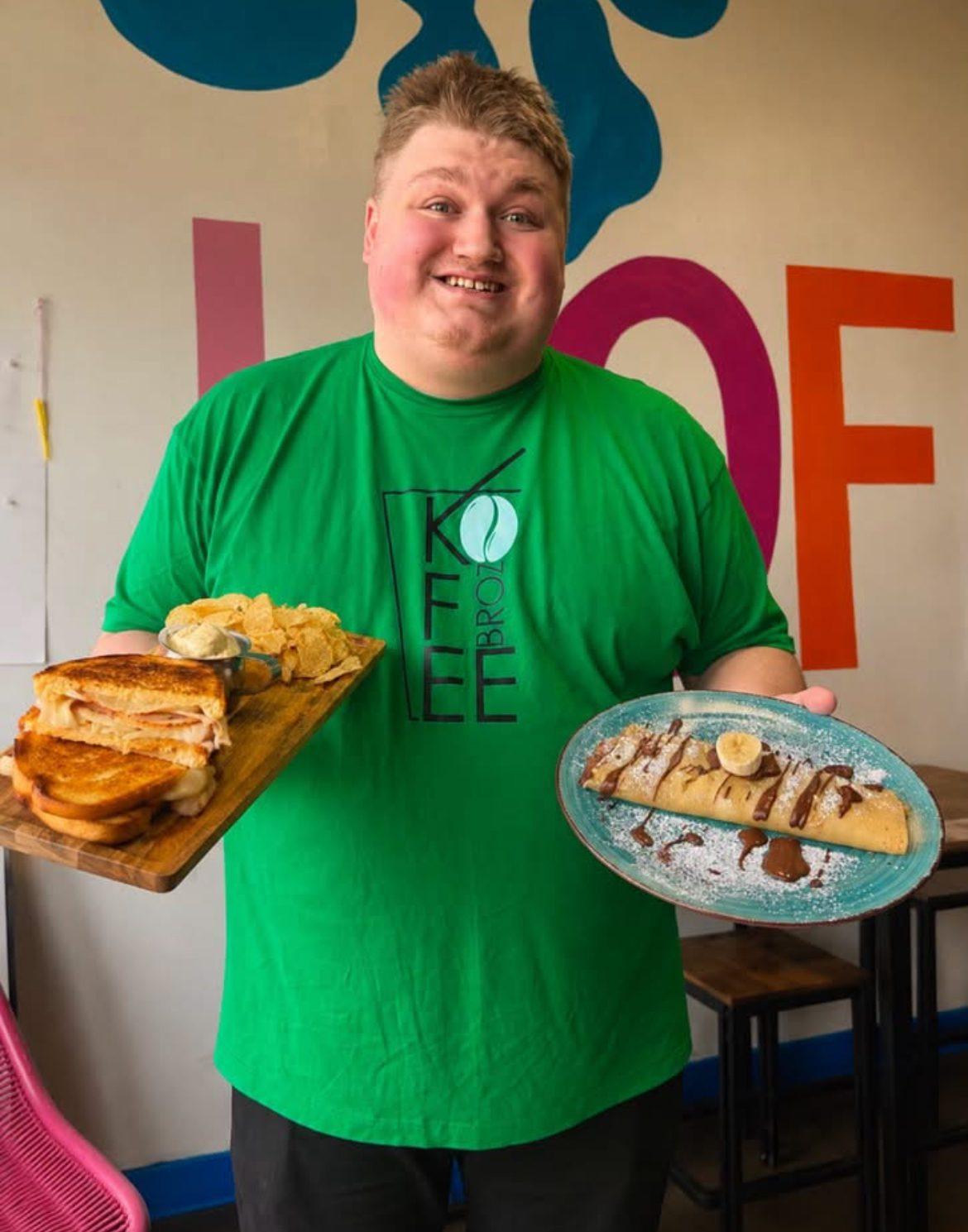 Kofee Broz team member in a Kofee Broz tee holding a sandwich-and-chips board and a crepe plate