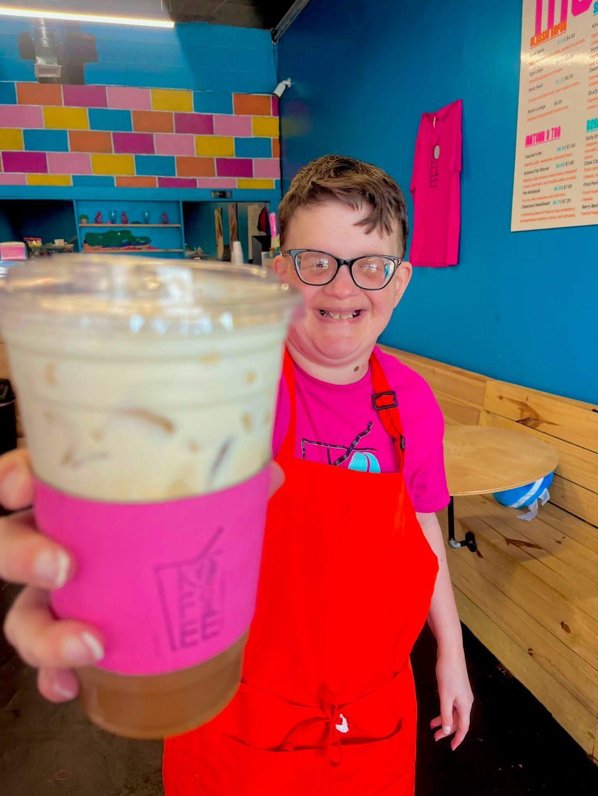 Kofee Broz team member in a red apron proudly holding up an iced coffee